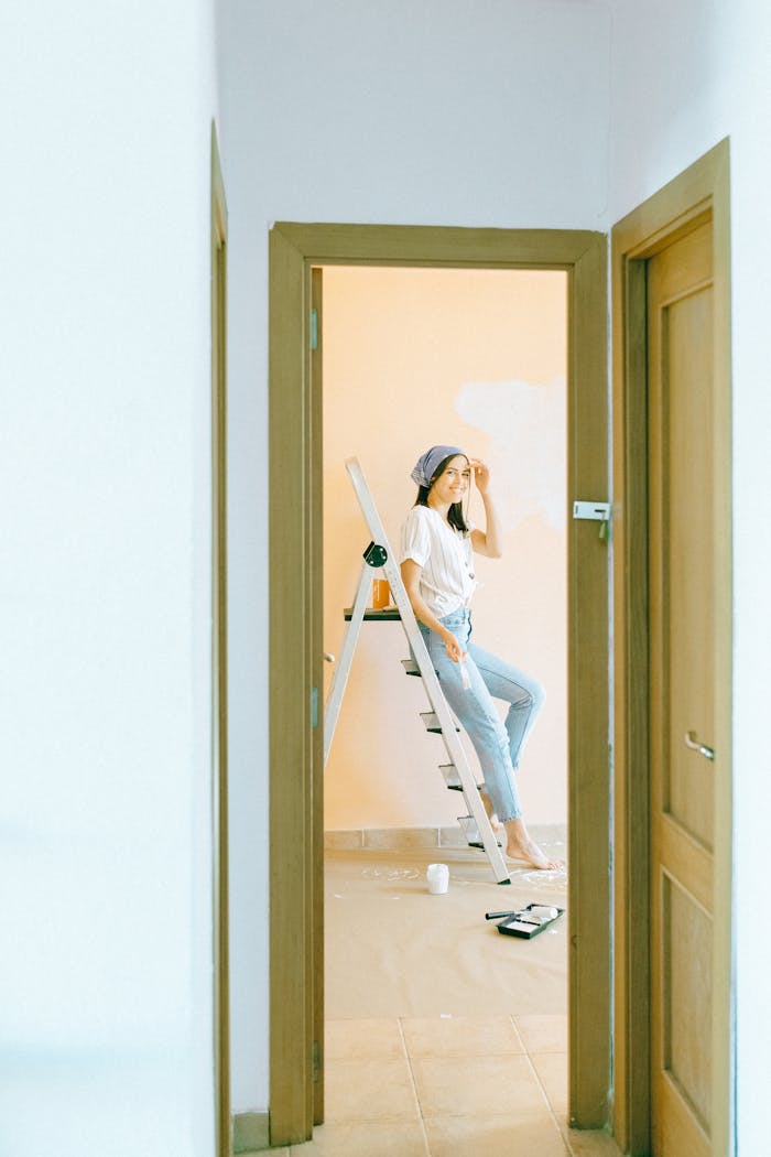 Woman sits on ladder painting a room with brush, bright interior setting.